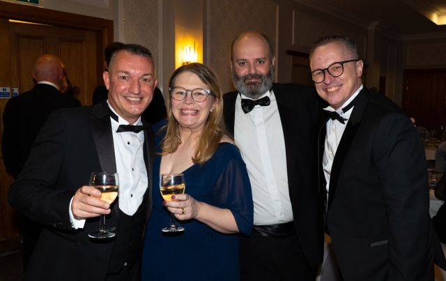 Four elegantly dressed individuals in tuxedos toast with glasses of wine at a formal event.